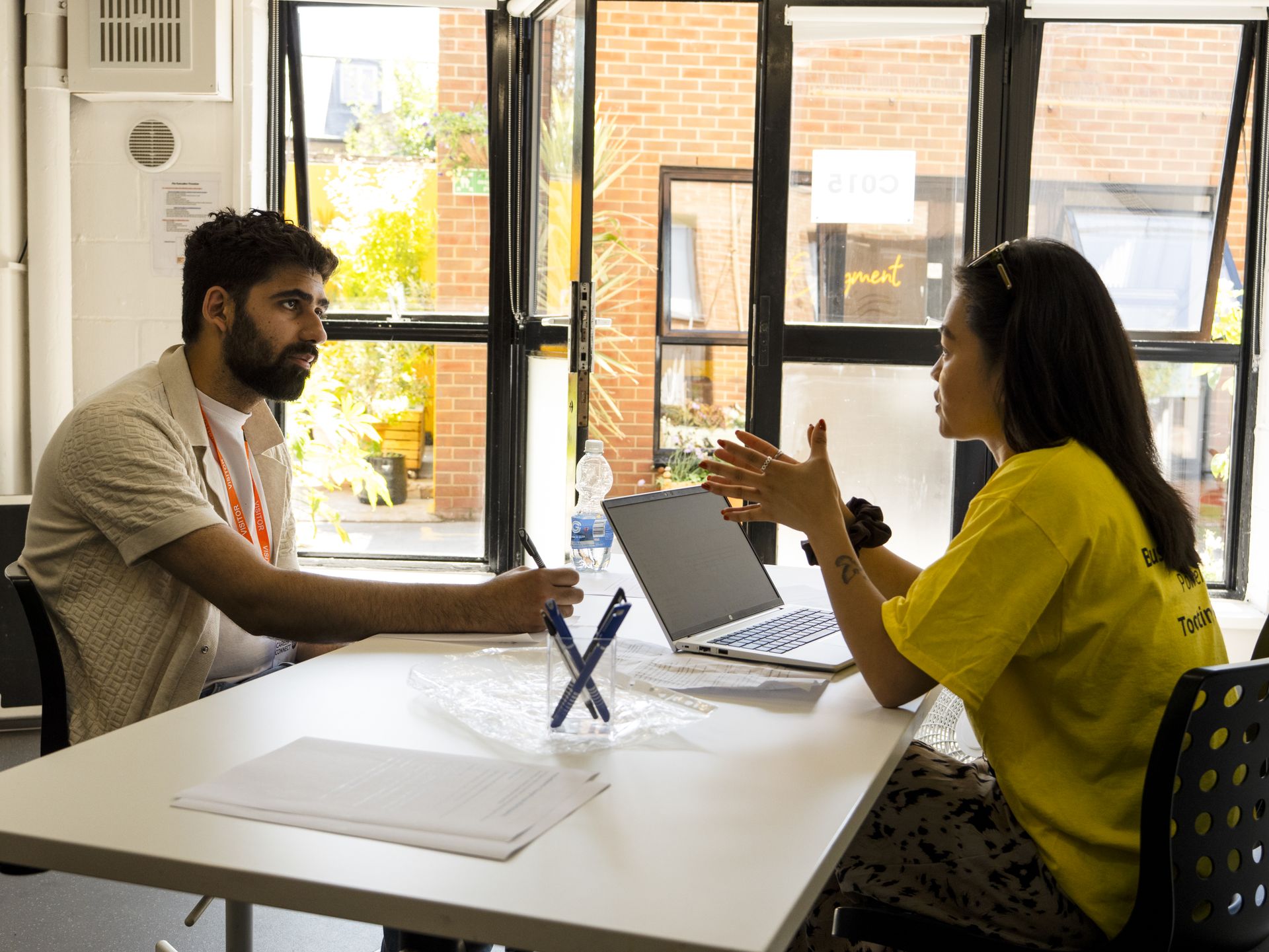 A woman in a bright yellow T-shirt is speaking to a man seated across from her at a table, during a one-to-one session indoors. She gestures while talking, with a laptop open in front of her. The man listens attentively, holding a pen. Sunlight streams through large windows behind them, revealing a brick courtyard outside. Career Connect Fest 2025 at Business Launchpad