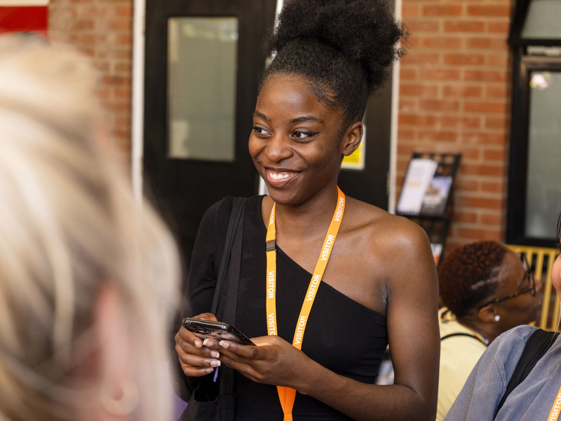 A young woman wearing a 'VISITOR' lanyard smiles while holding her phone during an indoor event. She is dressed in a black one-shoulder top and has a backpack over one shoulder. Other attendees and a brick wall are visible in the background. Career Connect Fest 2025 at Business Launchpad